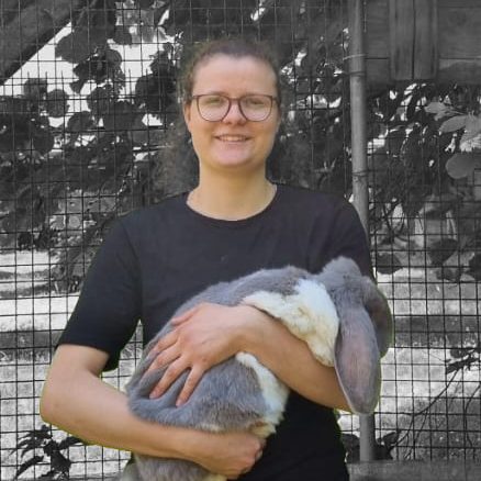 Cathryn Cyrus Frau mit Brille hält ein großes Kaninchen in den Armen, stehend vor einem Gitterzaun.
