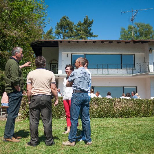 Gruppe von Menschen im Gespräch vor einem modernen Haus mit grünem Garten.